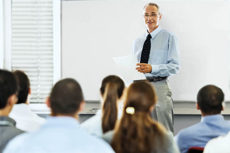 A male instructor in business attire lecturing a group of individuals.