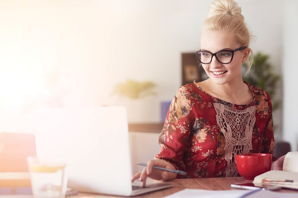 A woman in a red shirt and glasses smiling at her laptop screen.