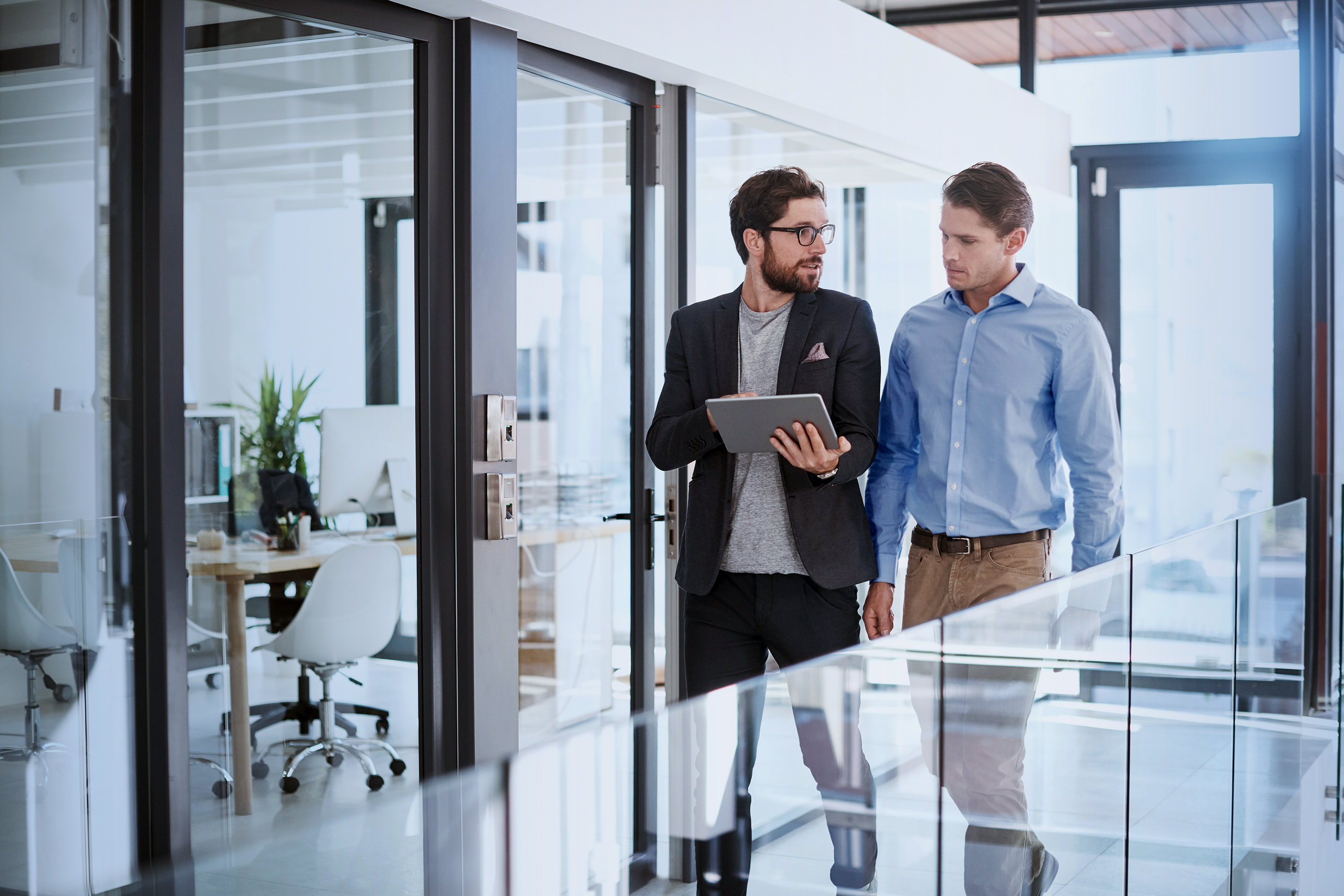 Two business professionals walking through an office corridor discussing what is on the tablet screen they are holding.
