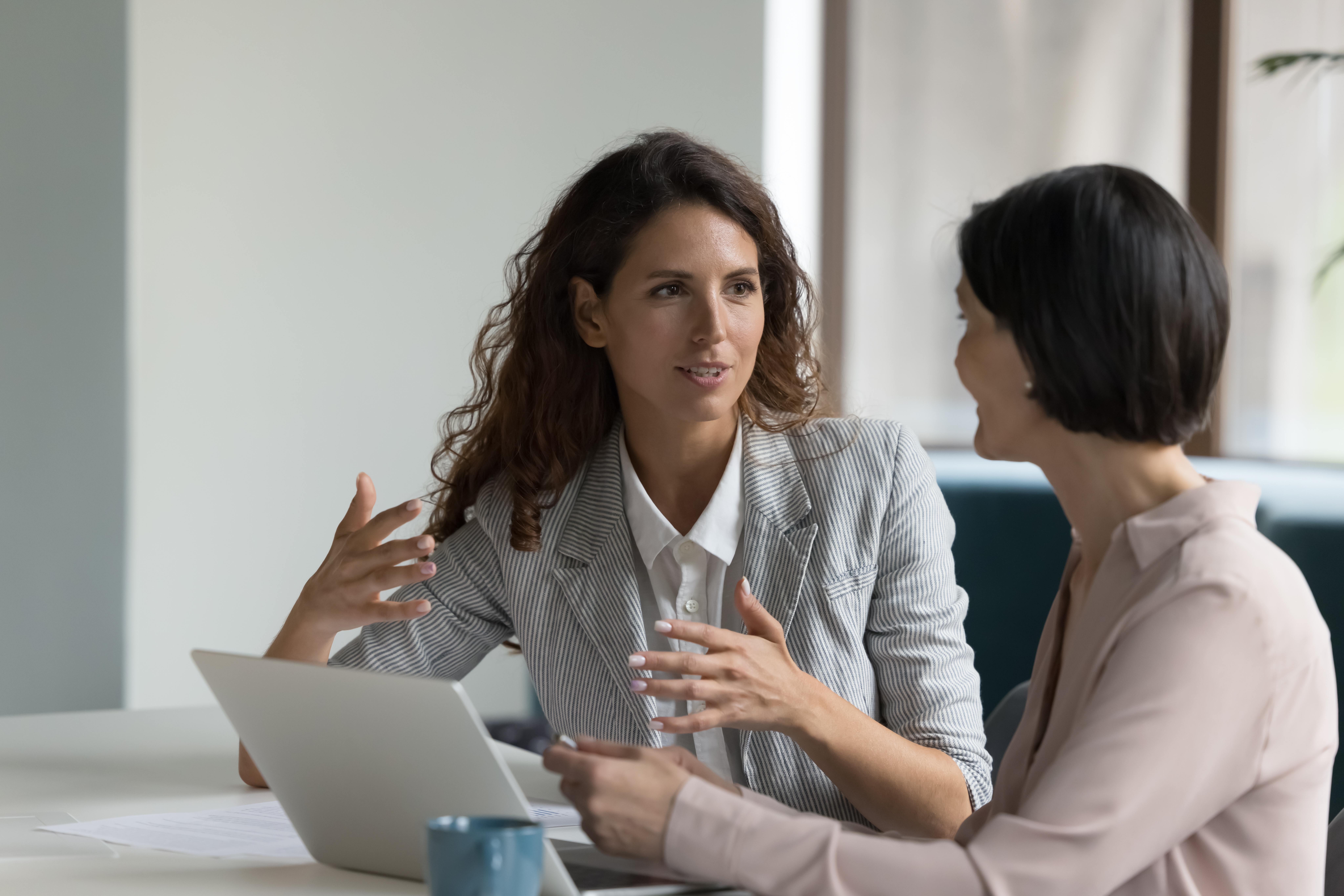 A career coach and a coachee, both dressed in professional business attire, sit at a conference table with a laptop open between them, actively engaging in a one-on-one coaching session as part of an outplacement program.